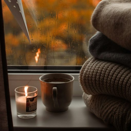 Window sill with a lit candle, a cup of tea and folded blankets, with orange autumn leaves seen through the rainy window