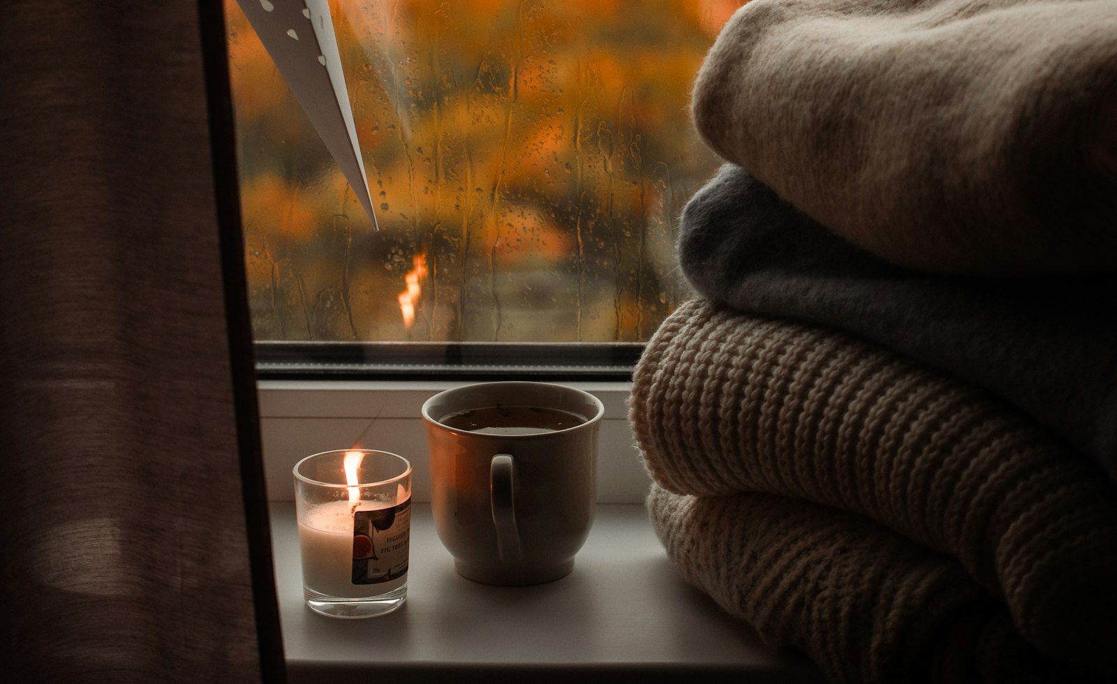 Window sill with a lit candle, a cup of tea and folded blankets, with orange autumn leaves seen through the rainy window